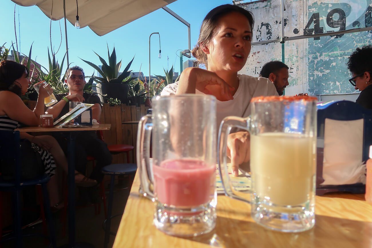 Close up of two jugs of pulque on rooftop with Kim talking in background