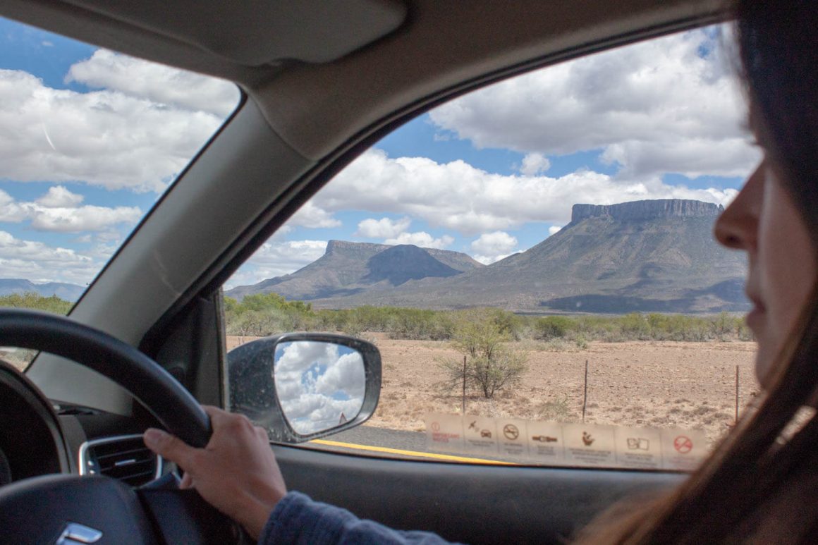 View through driver's side window of mountains, desert, and clouds in rearview mirror